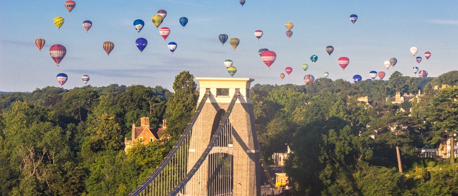 Clifton suspension bridge on a sunny afternoon with colourful hot air balloons in the sky above.